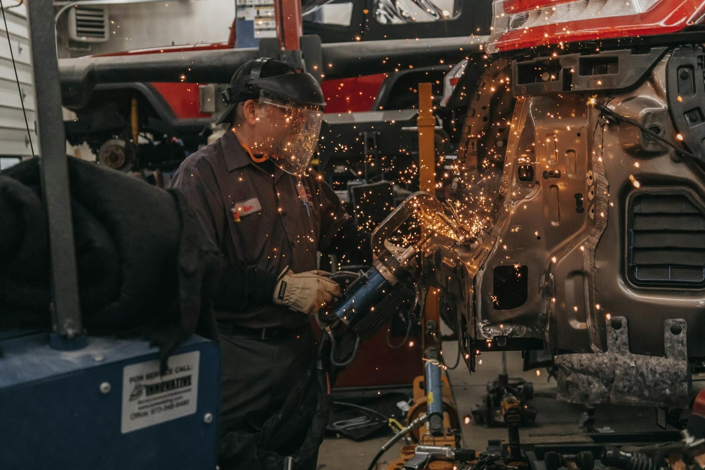An auto-body technician in a face shield grinding a stripped collision-damaged body shell, sparks flying, mid-repair